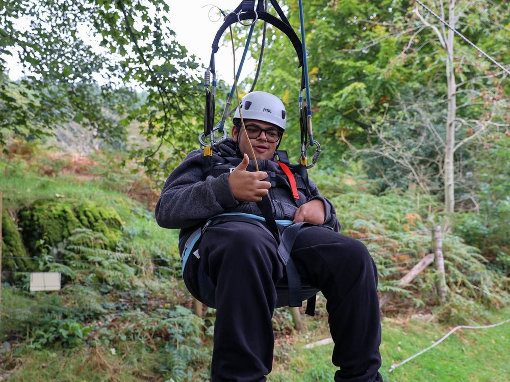 A young man wearing a crash helmet harnessed into a zipwire suspended in trees with his thumbs up