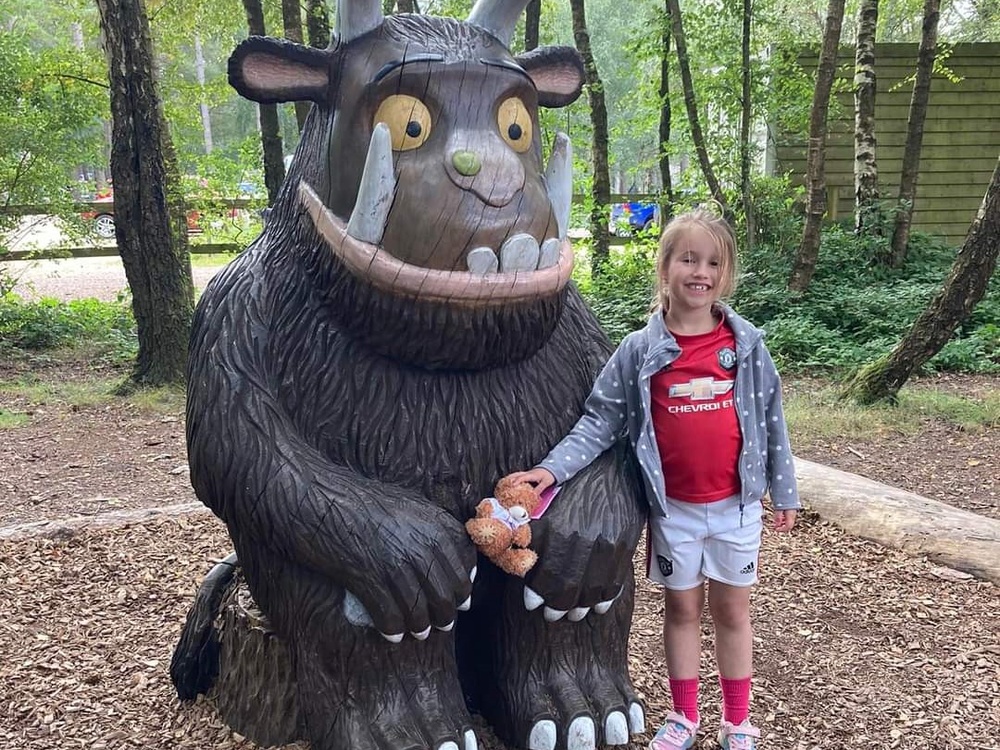 A young girl stands next to a large Gruffalo model in a forest, holding a Lily-branded teddy