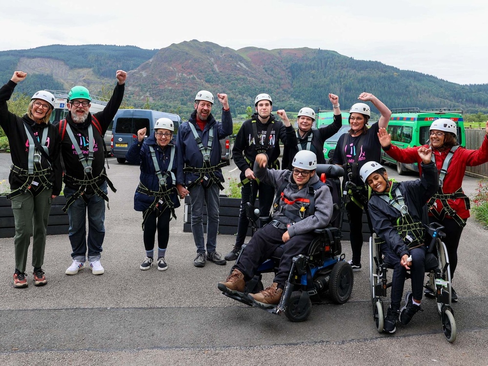 A group of people wearing outdoor clothing and harnesses and cheering, with hills in the background