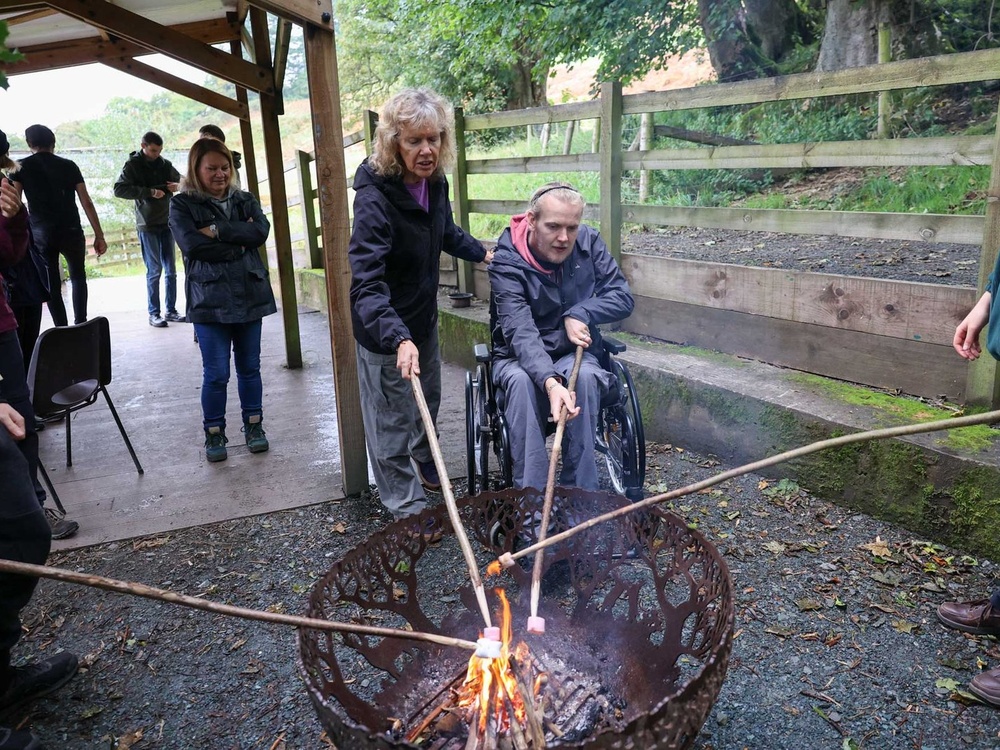A man in a wheelchair and a lady standing next to him are toasting marshmallows on sticks in a fire pit