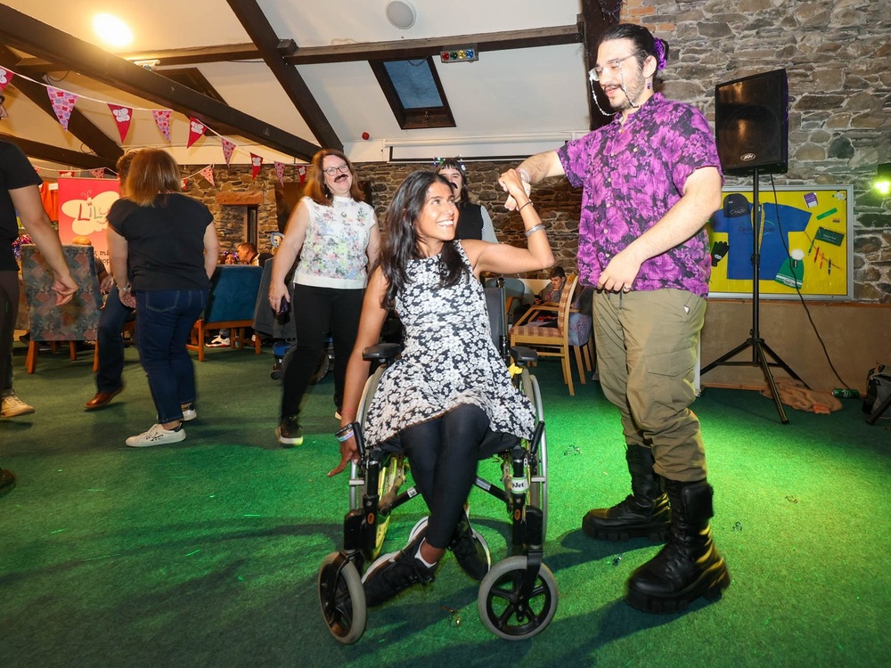A lady in a wheelchair is on a dancefloor holding the hand of a man who is dancing with her