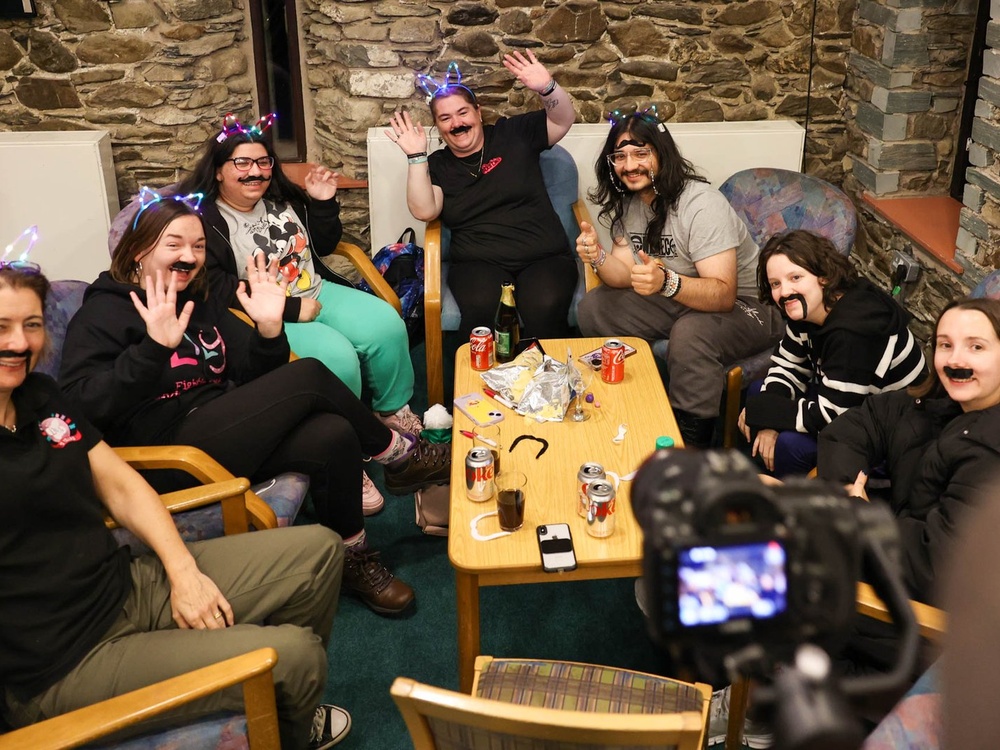 A group of people sitting around a low table laughing and wearing stick-on moustaches