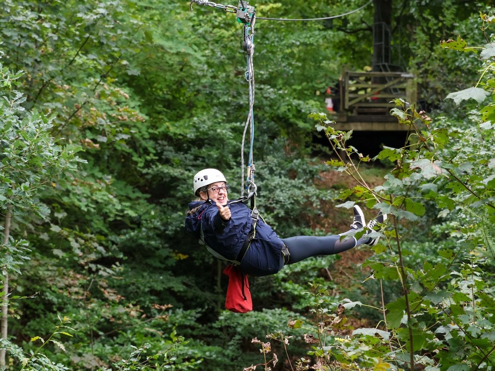 A lady dangles from a zip wire in trees giving a thumbs up to the camera