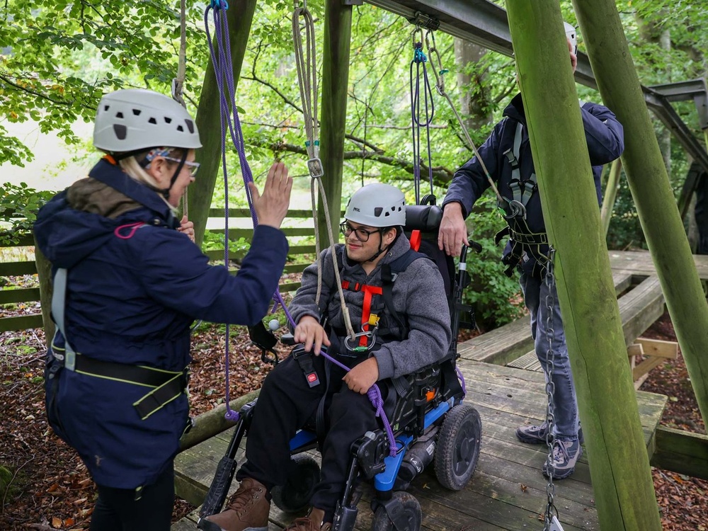 A man in a wheelchair reaches the end of a wooden treetop platform and high fives the lady waiting for him