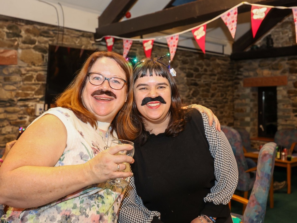 Two ladies with stick-on moustaches are laughing at the camera, one is holding a glass of wine