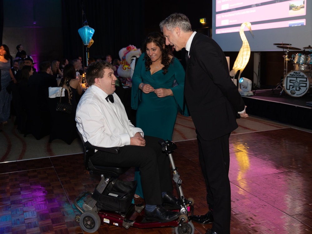 A young adult mito patient in a wheelchair talking to a man and lady on the dancefloor