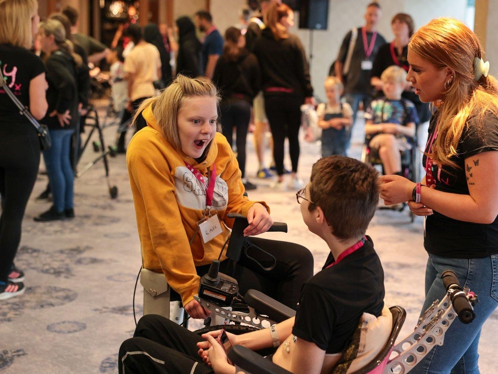 Two children, a girl and a boy, in wheelchairs are laughing and exclaiming together. Both are wearing Lily lanyards.