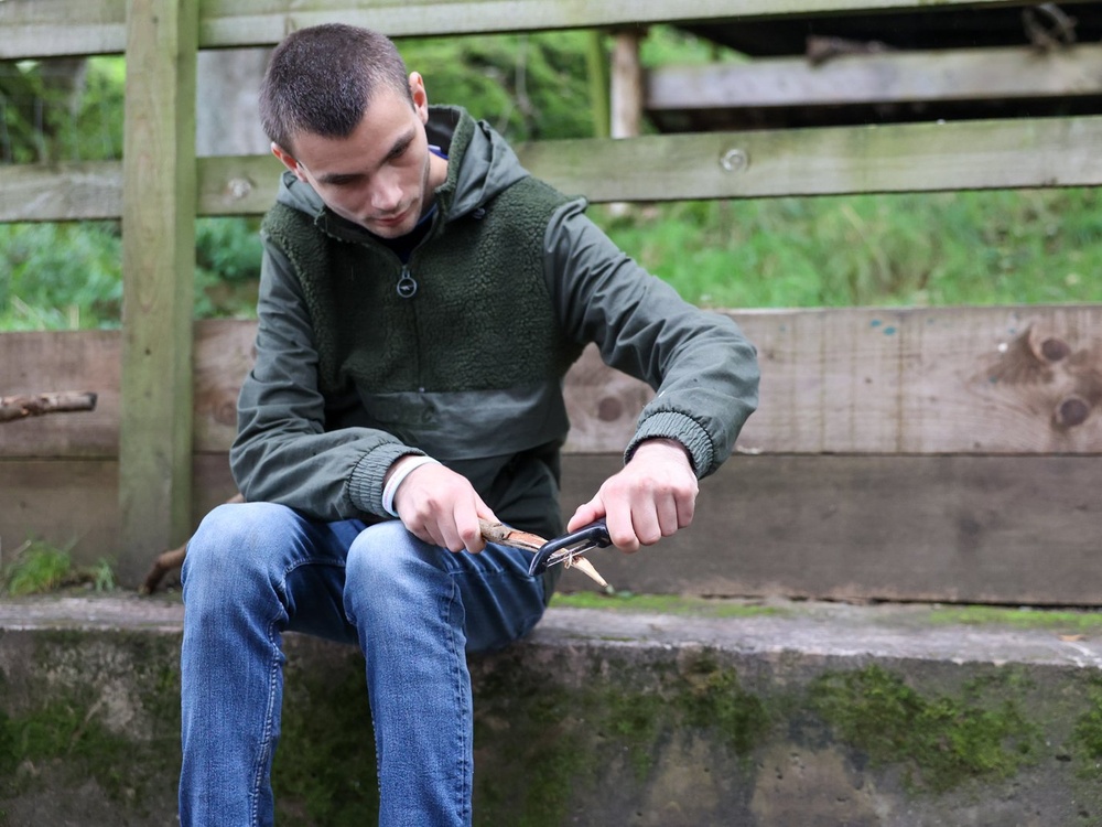 A young man is sitting outside with a look of concentration using a vegetable peeler to whittle a stick into a point