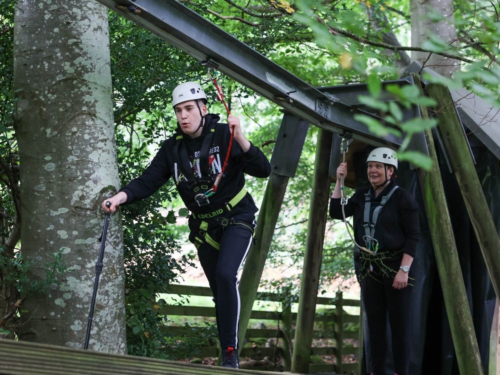 A young man in a harness with a stick is making his way across a wooden treetop platform