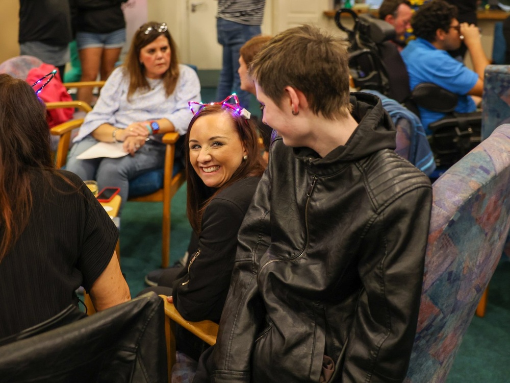 A lady in a chair is turning around to laugh with a young man sitting on the arm of the chair with his back to the camera