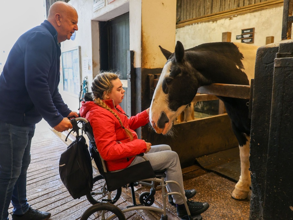 A young lady in a wheelchair strokes a horse's face in a stable
