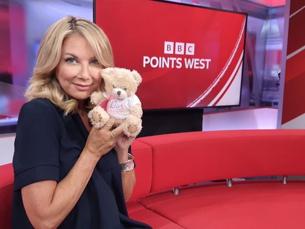 BBC presenter Alex Lovel in a TV studio holding a Lily-branded teddy and smiling