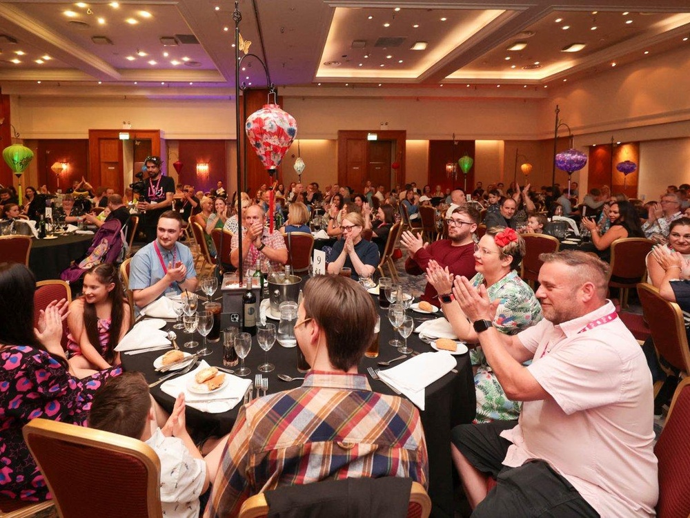 Many children and adults of all ages are seated around circular tables in a large room set up for a gala dinner, clapping