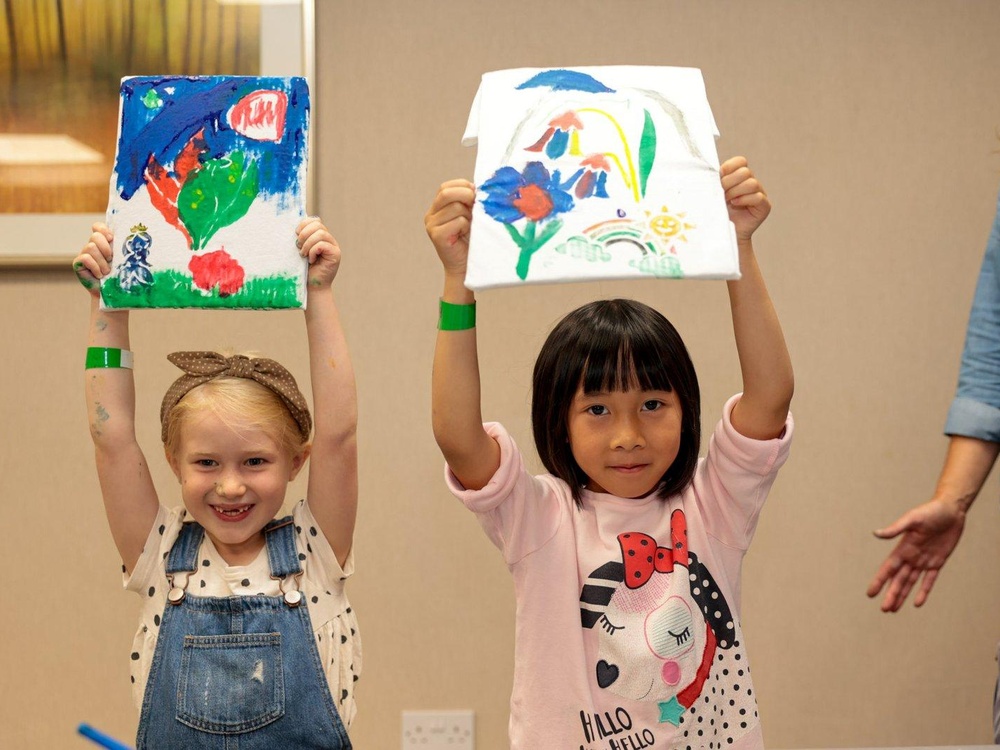 Two young girls are standing up holding paintings in the air above their heads and smiling
