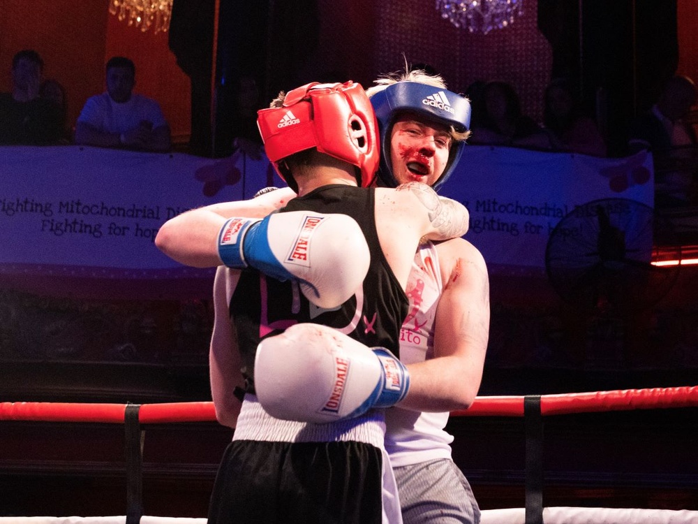 Two boxers, one with blood around his mouth, hug in the ring