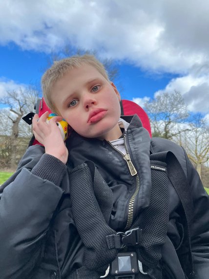 A blonde haired boy with mitochondrial disease looking into the camera holding a toy up to his right ear in his right hand, sitting in a an adapted buggy
