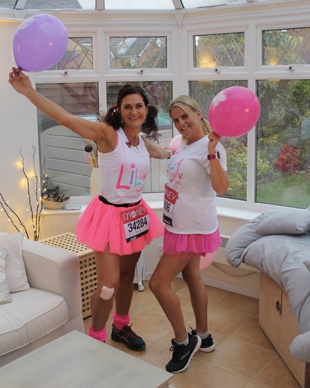 Two women smiling at the camera, one with dark hair and one with blond hair, both wearing Lily Foundation running tshirts and pink net short skirts with running numbers pinned to their fronts. The blond lady is holding a pink balloon and the dark haired lady a lilac balloon