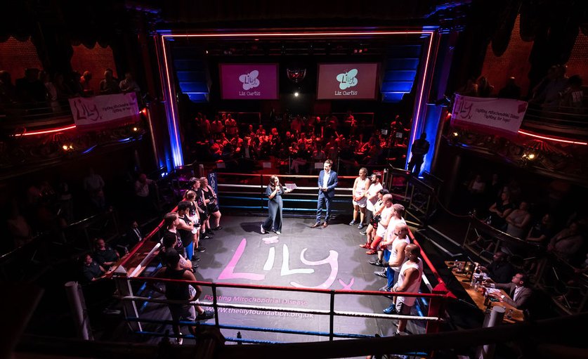 14 boxers stand around the edge of a boxing ring. The canvas has a large Lily logo in the middle. A woman is also in the ring talking into a micro phone. The room arounf them is full of people also listening to what the woman is saying