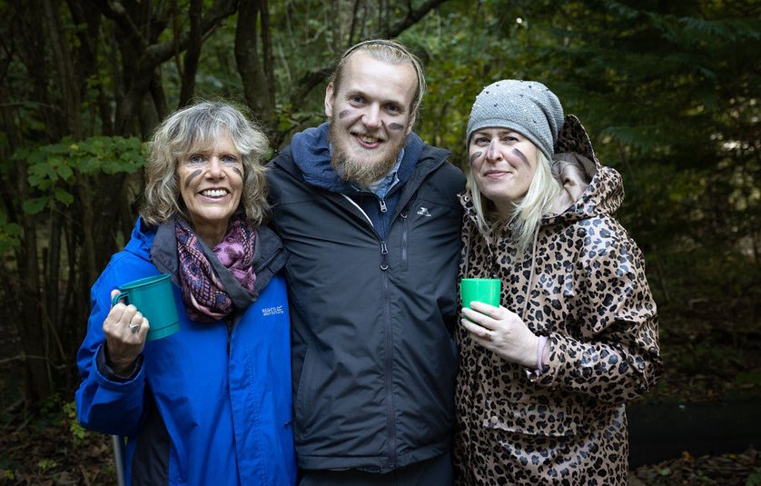 A man stands between two women. All three are dressed in outdoor clothes to keep them warm. They also have black make up stripes across their cheeks. The are all smiling into the camera