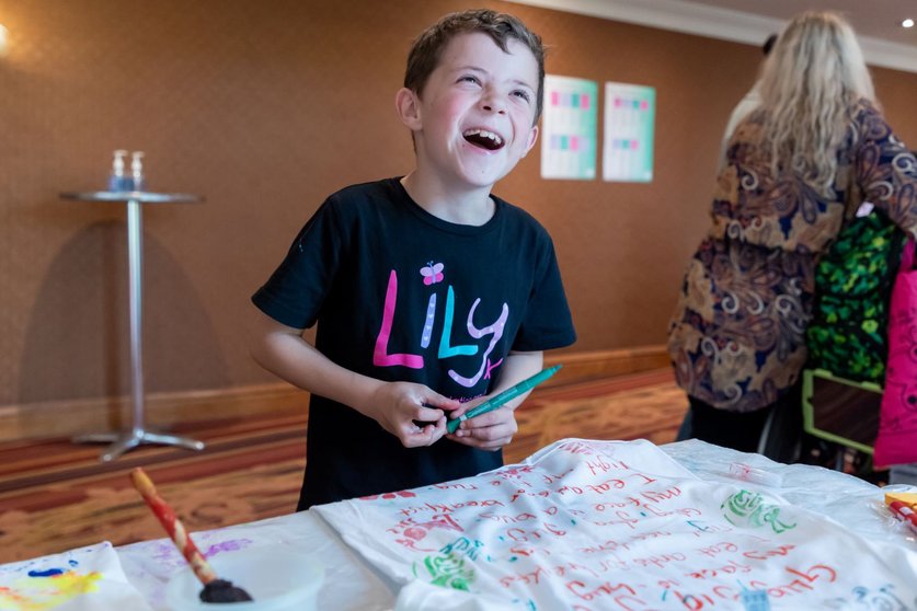 A little boy in a Lily Foundation Tshirt holds a pen. He is smiling and having fun