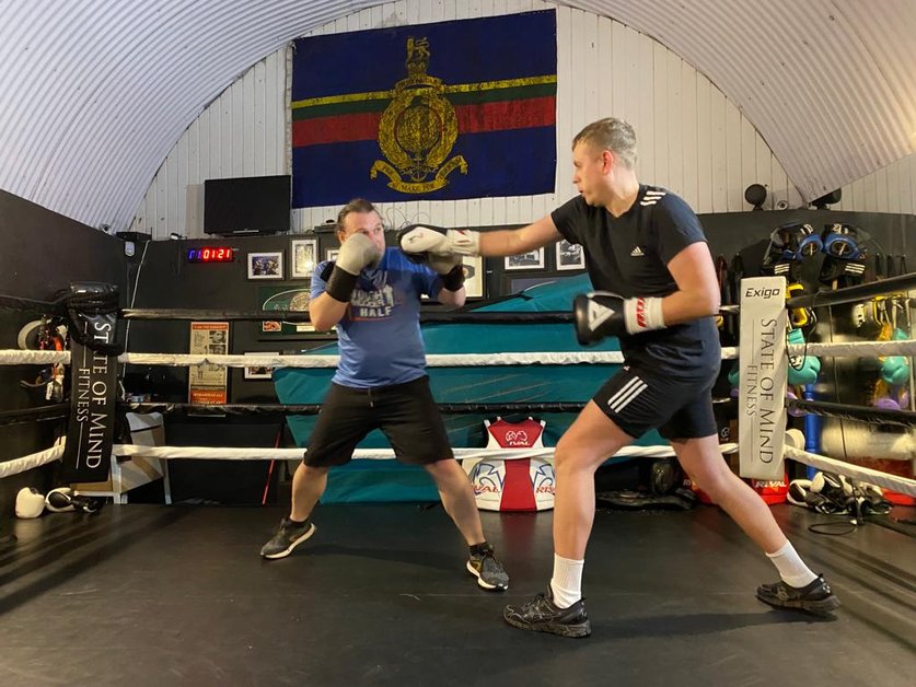 Two men boxing in a boxing ring. Both are wearing boxing gloves. One has his hands up guarding his face the other is swinging a punch towards him