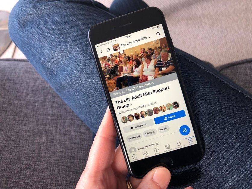 A hand is holding a smart phone. On it is a picture of a facebook page called The Lily Adult Mito Support Group with a picture of a group of people all sitting on chairs facing the same way looking in the same direction