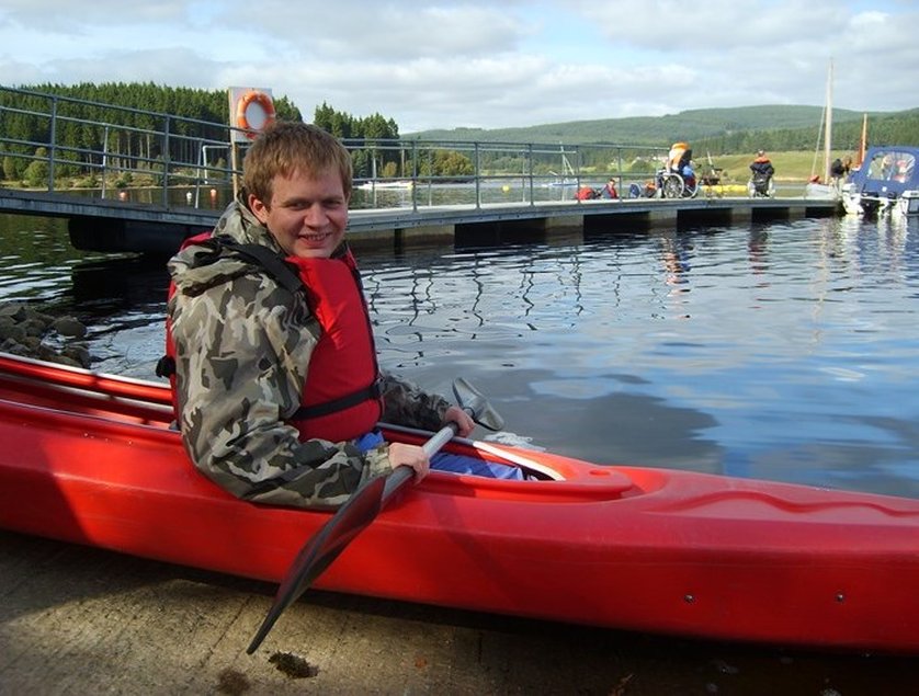 A smiling man looking at the camera in a red canoe holding a paddle on a concrete platform on the edge of a lake. Behind him is a pontoon with two people in wheel chairs near a a small boat 
