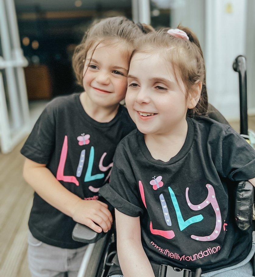 A little girl with mitochondrial disease in a wheel chair wearing a Lily Foundation tshirt. Stood beside her is a younger little girl holding onto the arm of the wheel chair. She is also wearing a Lily Foundation T-shirt