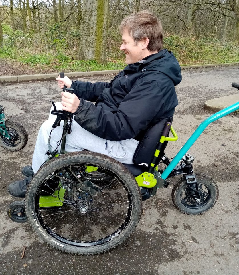 A side on view of a smiling man in a muddy Mountain trike using the its control levers 