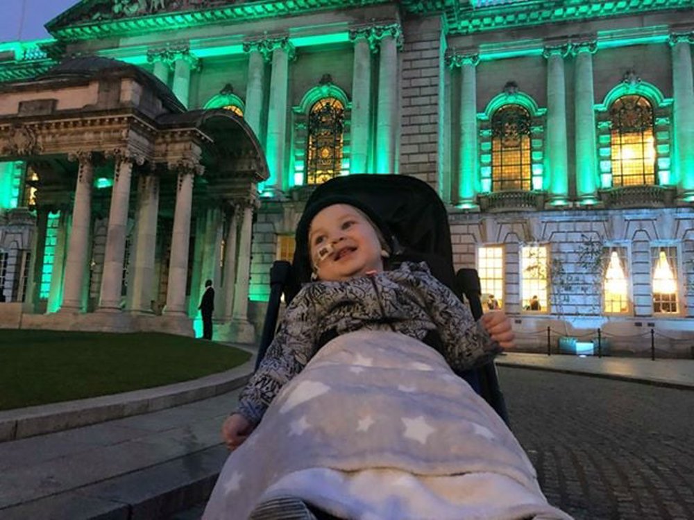 A child in a buggie with a feeding tube in their nose Infront of a building lit up green