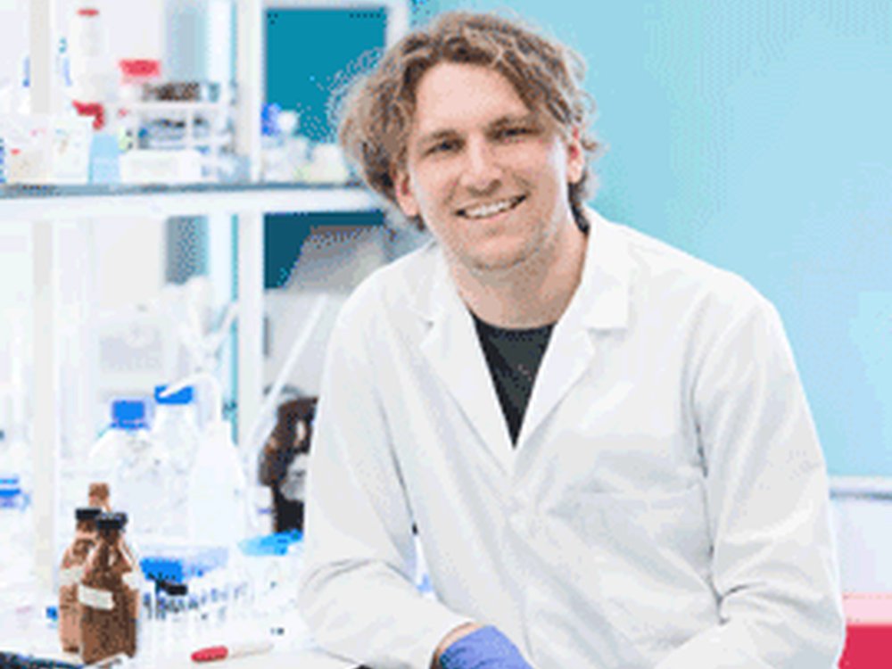 A man smiles at the camera he is wearing a white lab coat and blue medical gloves and leaning on a laboratory bench