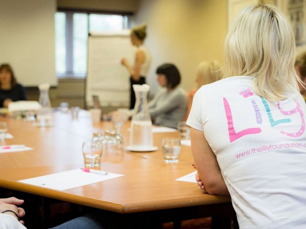 A group of adults sat around a large table all looking at a woman stood in front of a flip chart holding a pen