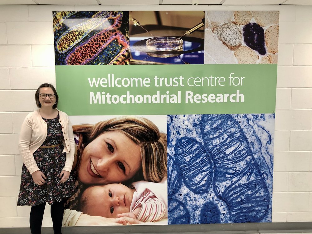 Katie Waller, standing in front of a huge sign for The Wellcome Trust Centre for Mitochondrial Research