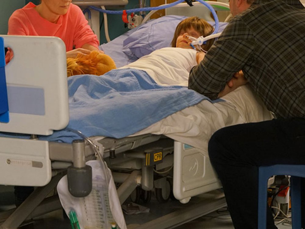 Two men and a woman sit beside a hospital bed. In it is a little boy who is asleep with a breathing tube