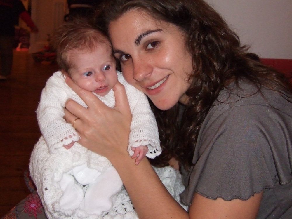 A lady with long dark hair cuddling a very young baby wrapped in a white knitted dress