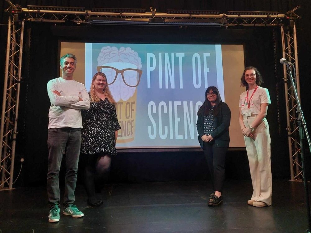 Four people stand in front of a large screen that says Pint of Science