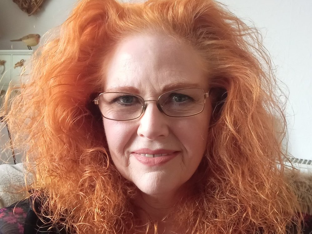 A head shot of a smiling woman with orange hair and glasses