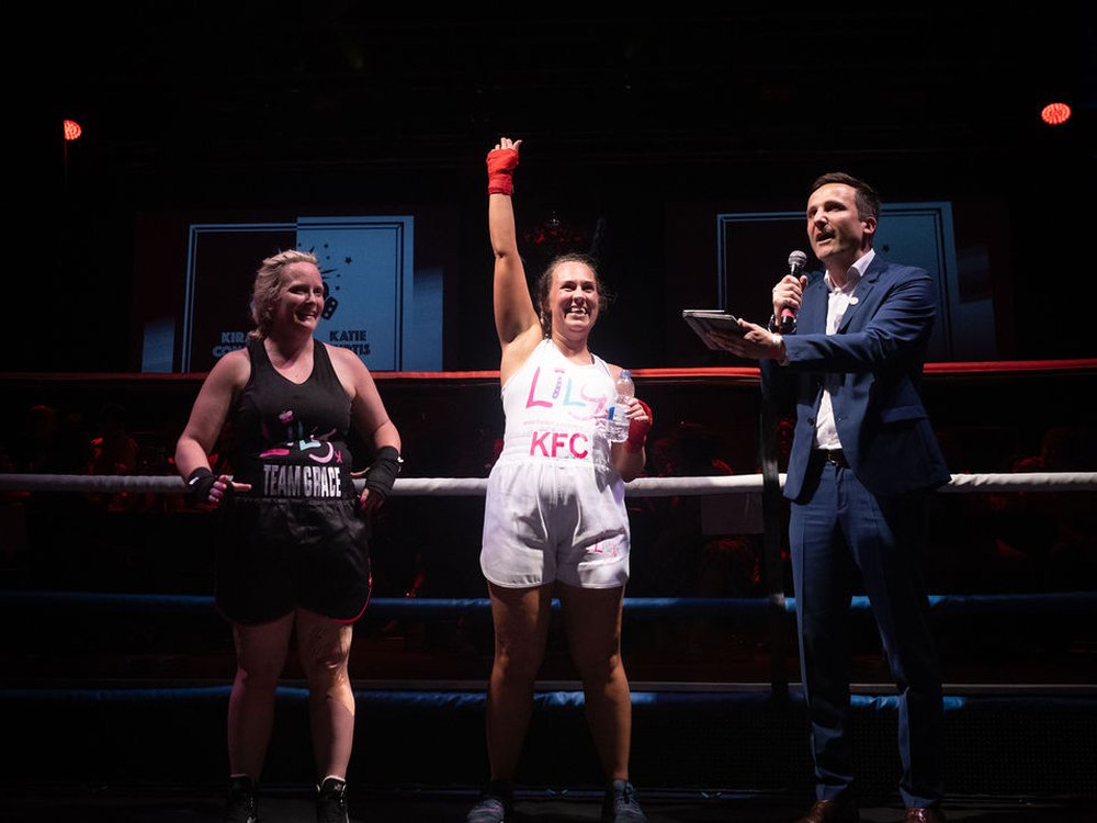 two women stand in a boxing ring one is holding her arm up in the air next to a man talking into a microphone