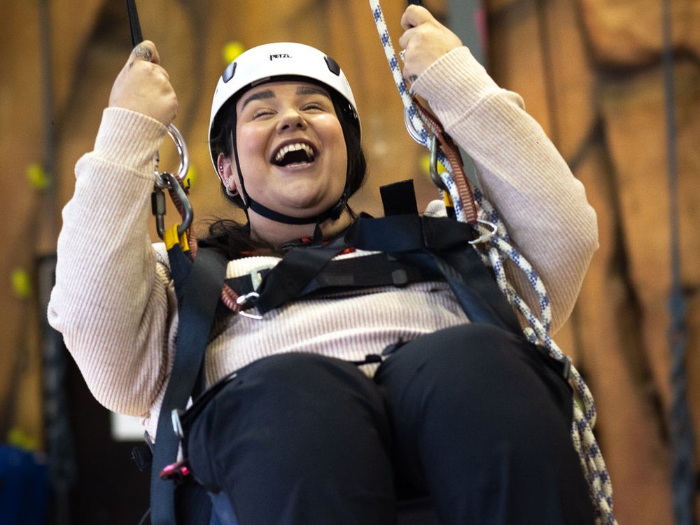 A smiling woman in front of a climbing wall in a specially adapted harness