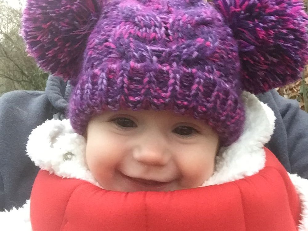 A little girl in a purple woollen hat with puple boobles each side smiles into the camera