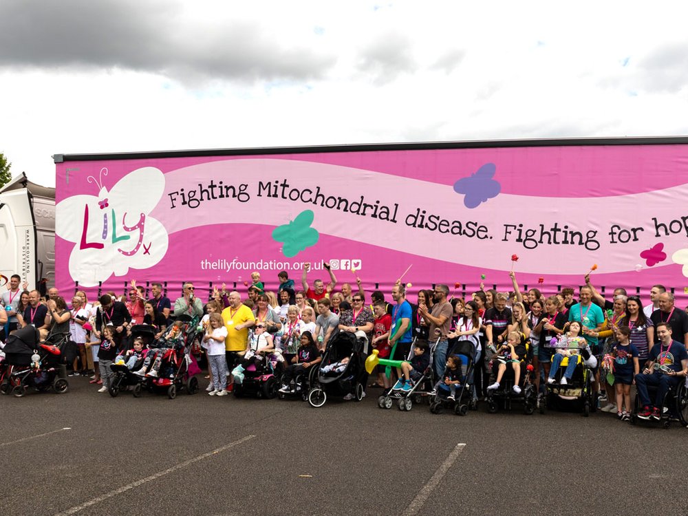 A large group of adults and children some with mitochondrial disease stand along the side of a large lorry with a Lily logo