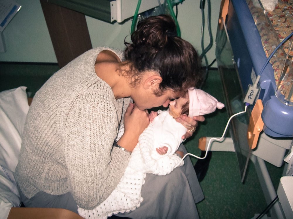 A Women sat in a chair holding  a small baby. They are touching noses. The baby is attached by wires to a medical device