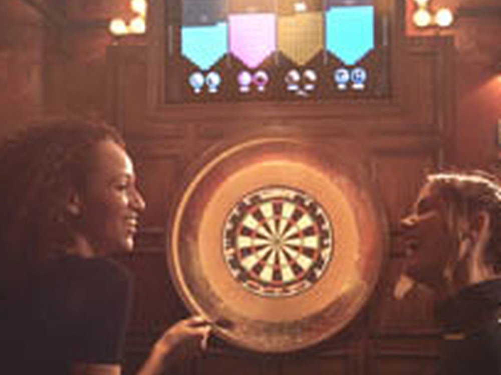 Two women stand either side of a lit up dart board looking at each other and smiling