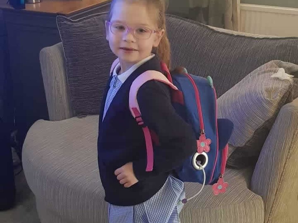 A young girl in a school uniform wearing a backpack with feeding tube equipment