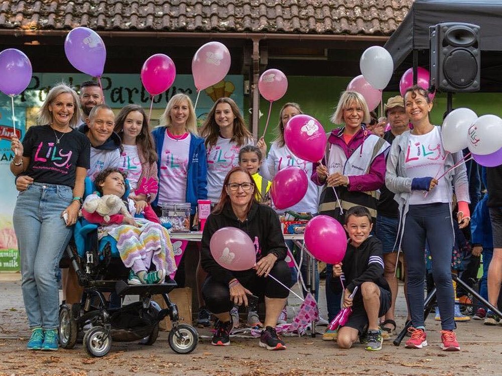 Group of people sitting and standing together wearing Lily Foundation tops and holding pink and purple balloons