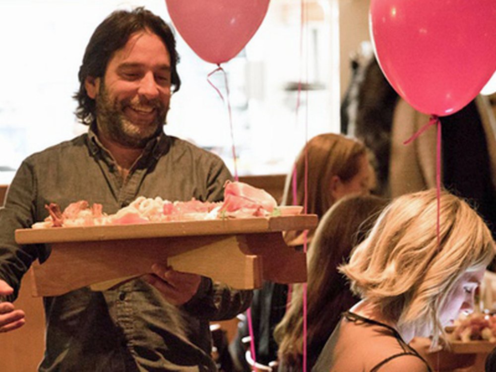 A man with shoulder length dark hair in a restaurant carrying a platter of food with pink Lily balloons in the background