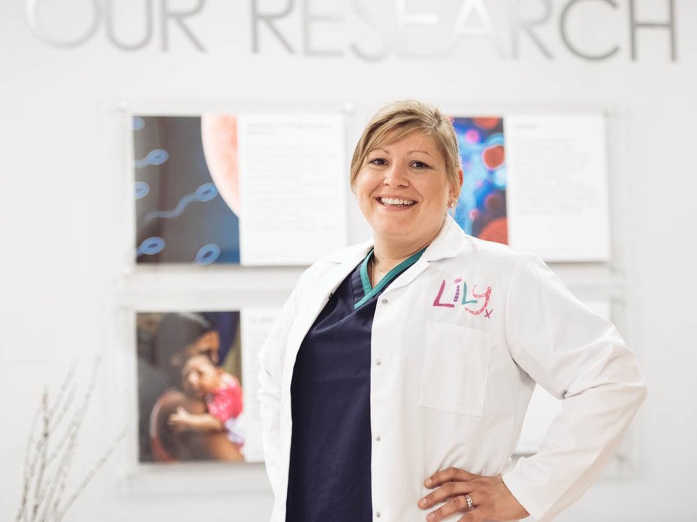 A lady in a white lab coat with a Lily Foundation logo smiling at the camera with her hands on her hips in a lab