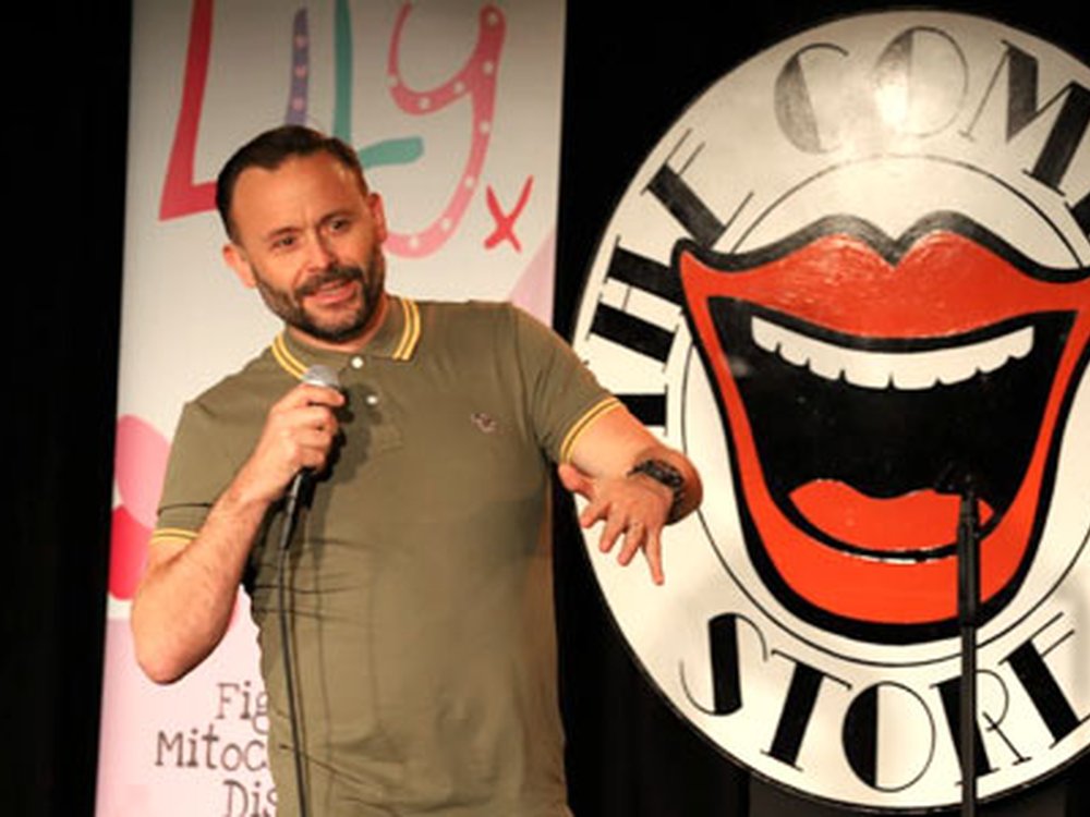 Comedian Geoff Norcott standing in front of the Comedy Store logo holding a microphone at the Lily comedy night