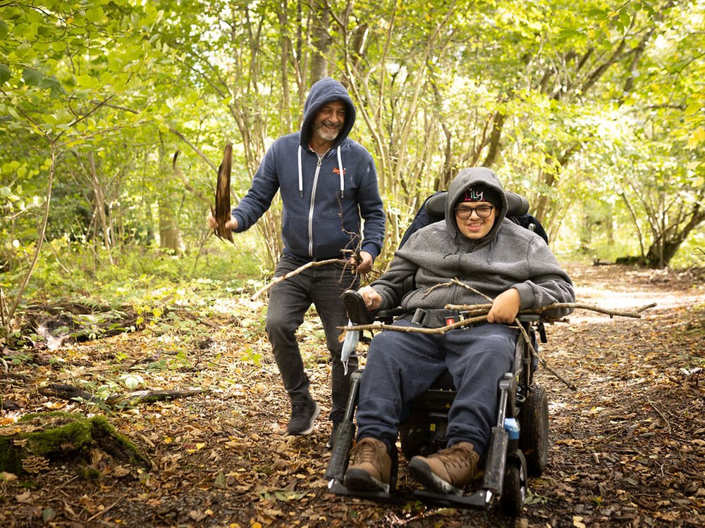 A young adult mito warrior in a wheelchair with his carer in trees at the Lily Foundation young adult weekend