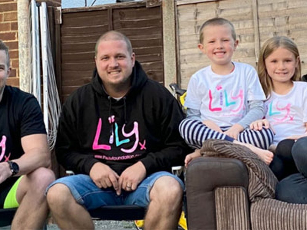 Three adults and two children wearing Lily Foundation tops and sitting together in front of a fence